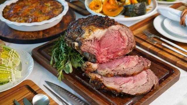 A large, well-cooked roast beef on a wooden cutting board is the centerpiece of a festive meal. Surrounding it are a variety of dishes, including a baked potato, a casserole, and a salad. The table is set with utensils and a white tablecloth, creating a warm and inviting atmosphere.