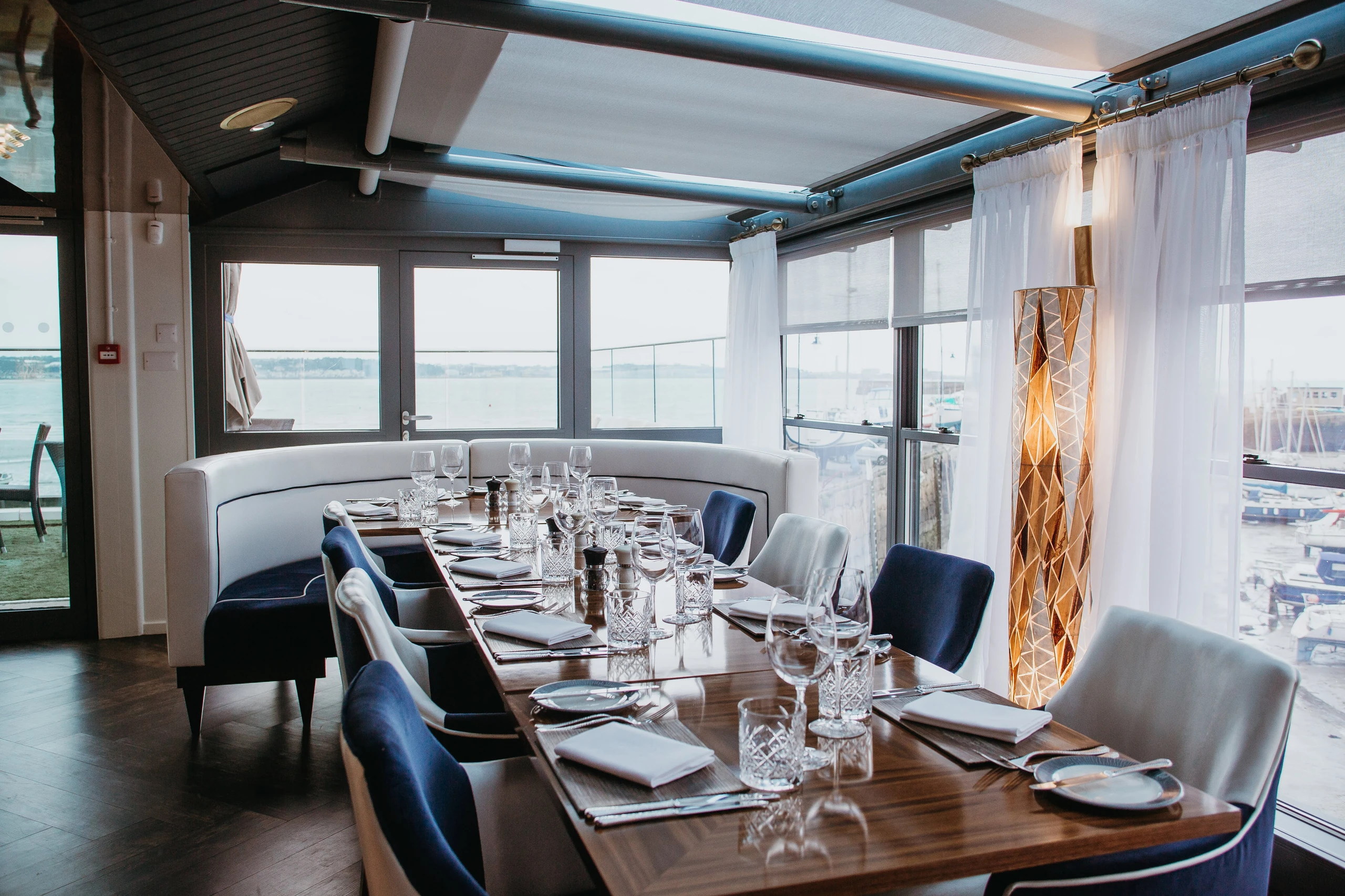 The image shows a modern dining room with a wooden table, white chairs, and blue cushions. The table is set with silverware, glasses, and napkins. A large window offers a view of the ocean, and a white sofa is in the background. A tall lamp with a orange and white shade stands on the right side, casting a warm glow. The room is well-lit and has a modern aesthetic.