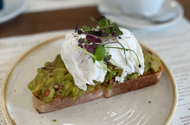 A white plate holds a slice of toast with guacamole, poached egg, and garnish, placed on a white tablecloth. The toast is topped with a creamy guacamole spread, and the egg is poached and garnished with red pepper flakes. The background is blurred, focusing on the plate and its contents.