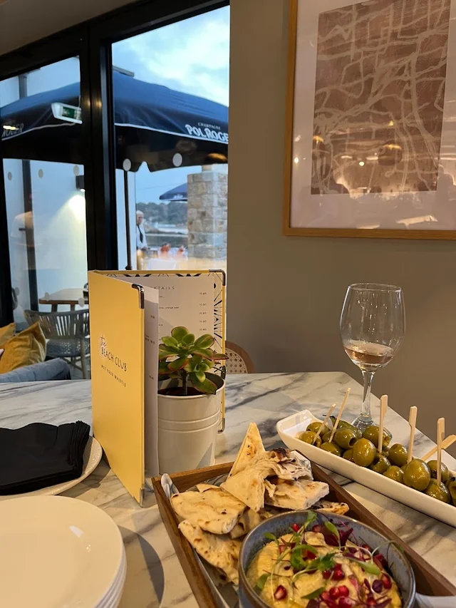 The image shows a dining table set for a meal, with a tray of olives, a glass of wine, and a bowl of salad. The table is near a window with a view of a patio, and a potted plant adds greenery. The scene is inviting and elegant.
