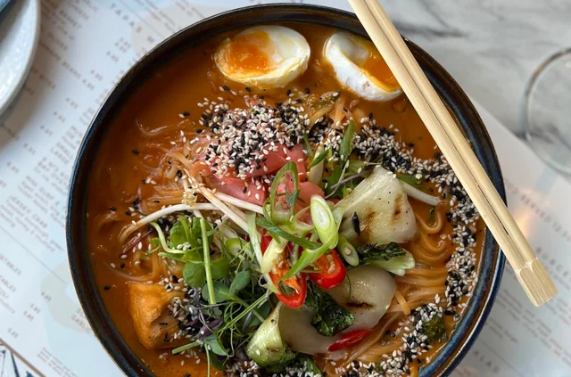 A black bowl filled with ramen noodles, vegetables, and an egg is on a white tablecloth with a menu. Chopsticks are nearby, and a glass of water is in the background. The scene is visually appealing and inviting.
