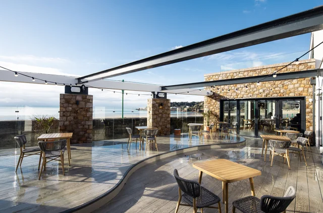 The image features a modern outdoor patio with a curved wooden floor, surrounded by stone pillars and a large glass wall. Tables and chairs are arranged for guests, and a stone wall with a bar and bar stools is visible. The patio overlooks a serene ocean view, creating a tranquil atmosphere.