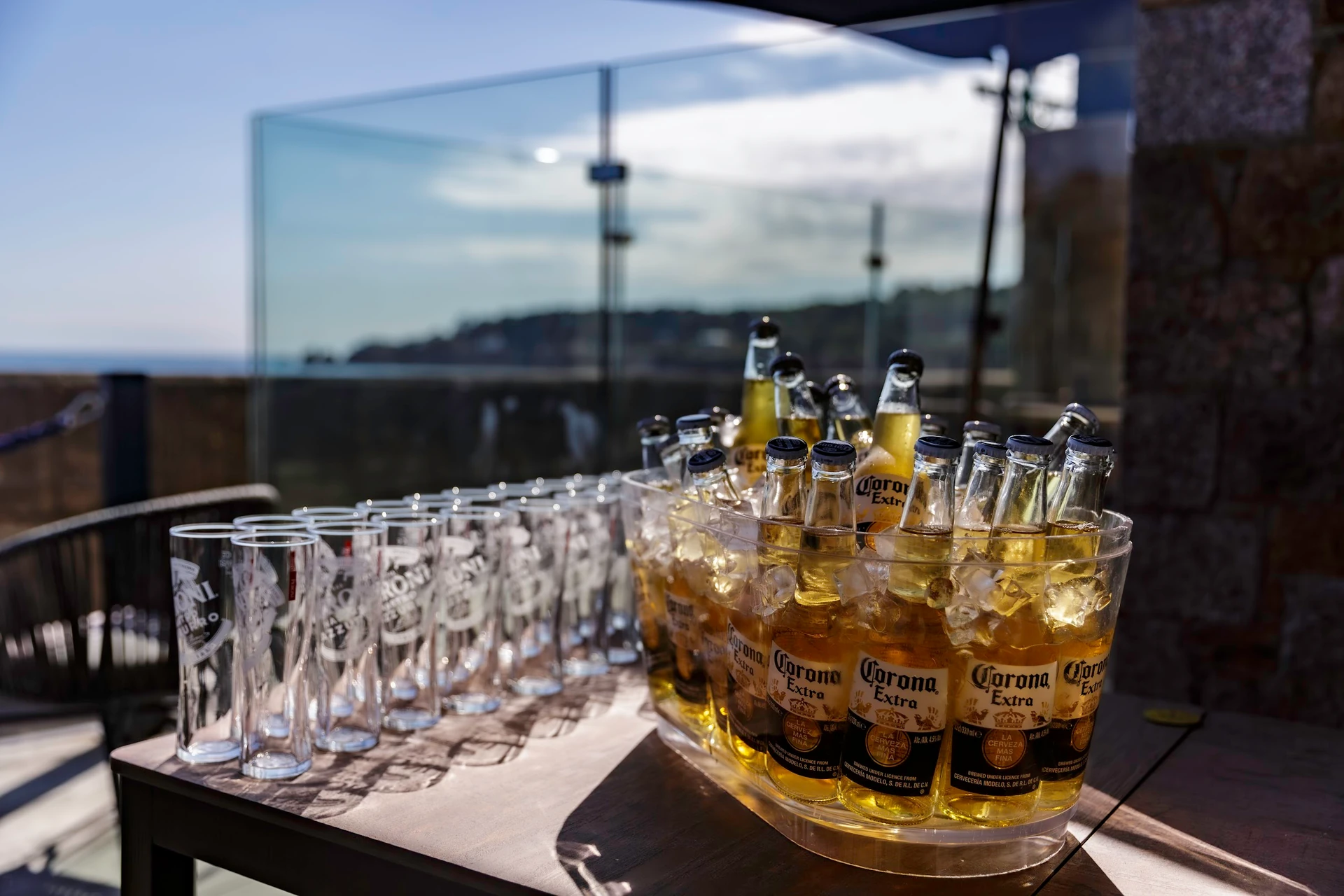 A serene outdoor setting features a wooden table with a glass bowl filled with beer glasses, arranged in a semi-circle. The glass bowl has "Braúna de la Batalha" text, indicating the brand. The table is on a balcony overlooking a scenic view of a mountain range and a clear blue sky. The scene is tranquil and inviting, with the beer glasses ready for a social gathering.