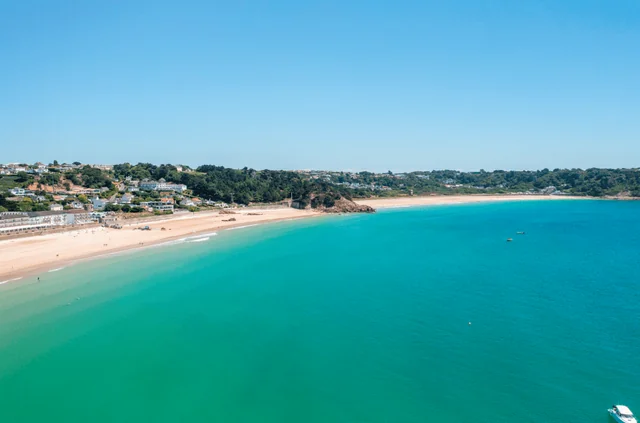 The image features a serene beach scene with a sandy shore, turquoise water, and a few boats. The beach is surrounded by a hill with houses and trees, and the sky is clear blue. The perspective is from above, offering a comprehensive view of the beach, water, and hill. The image is a beautiful representation of a typical day at the beach.