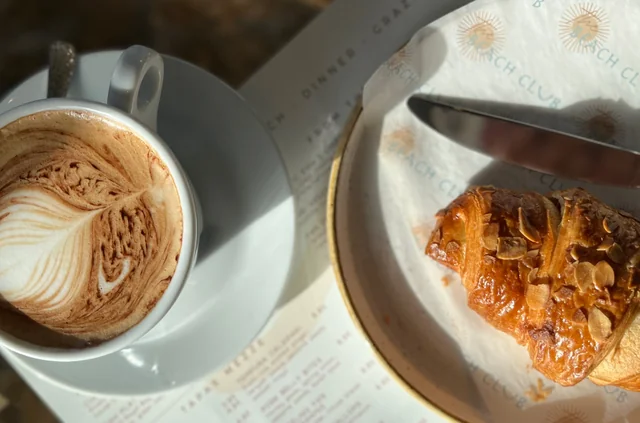 A serene breakfast scene features a white coffee cup with latte art on a white saucer, next to a golden croissant on a white plate. The coffee cup is on a white napkin with a blue pattern, while the croissant is on a white plate with a blue pattern. The objects are arranged in a balanced manner, creating a harmonious composition.