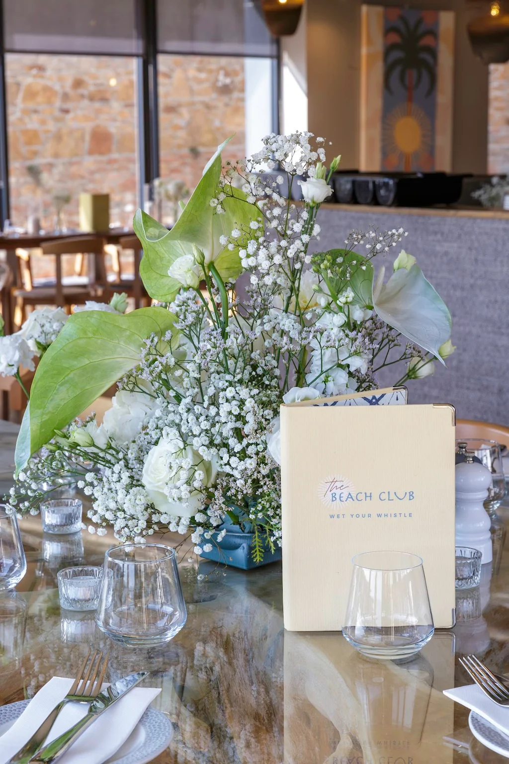 The image features a beautifully set table with a blue vase of white flowers, a vase of green leaves, and a white napkin. The table is set with silverware and glasses, and a menu is visible in the background. The scene is set against a rustic stone wall and a window, creating a warm and inviting atmosphere.