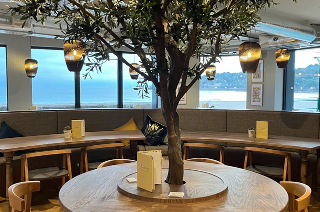 The image features a modern restaurant with a wooden table surrounded by chairs, a large tree with a blue vase and white candles, and a window offering a view of the ocean. The table is positioned in the center, with chairs on either side, and the tree is centrally located. The room is illuminated by hanging lamps and a large window with a view of the ocean. The arrangement of objects creates a harmonious and inviting atmosphere.