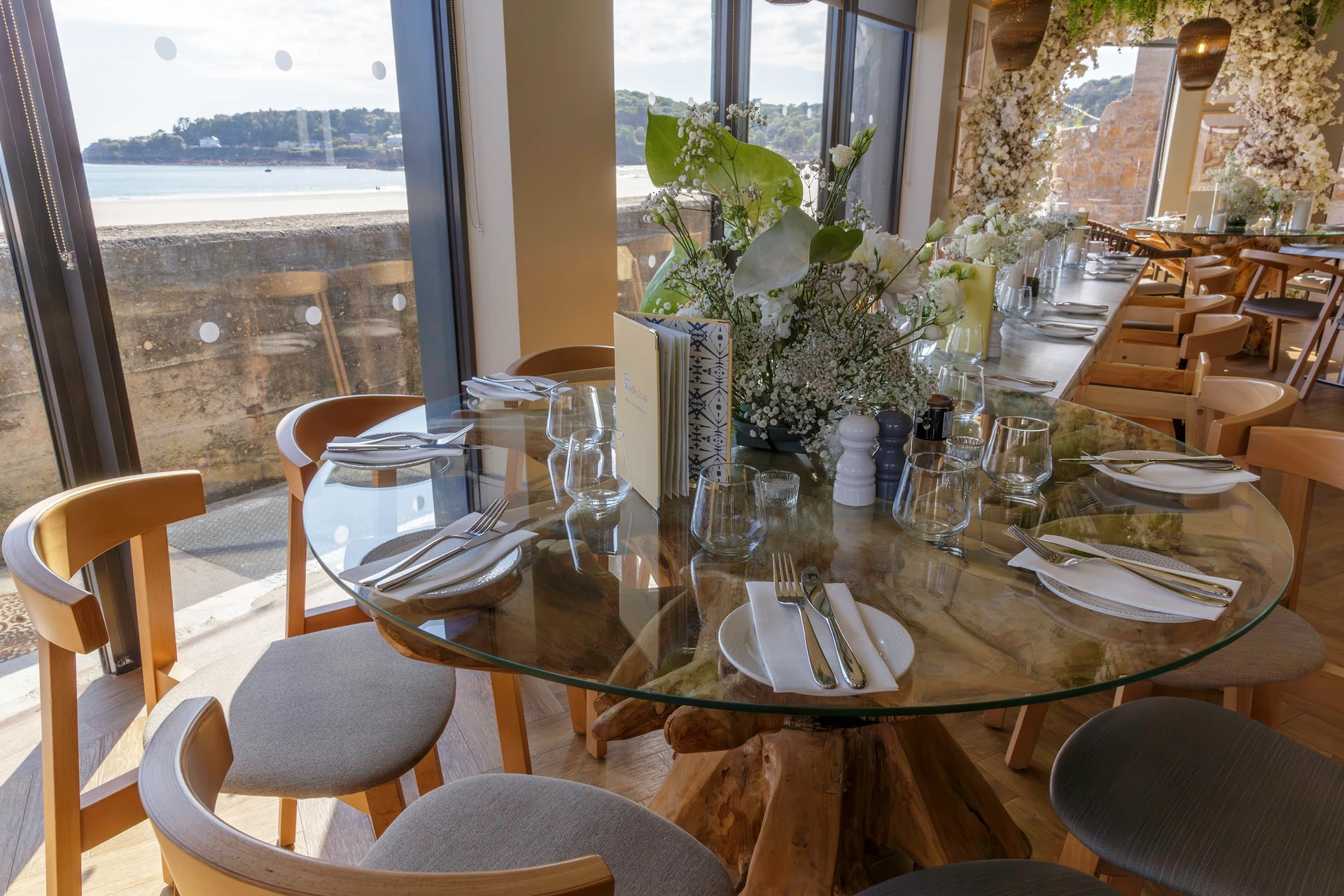 A modern restaurant with a glass table set for six, surrounded by wooden chairs, features a vase of white flowers and greenery, a vase of white flowers, and silverware. The table is set for a meal, with a vase of white flowers and greenery in the center. The restaurant has a serene ocean view through large windows, and the floor is made of wood. The scene is well-arranged and inviting, with a focus on the table setting and the ocean view.