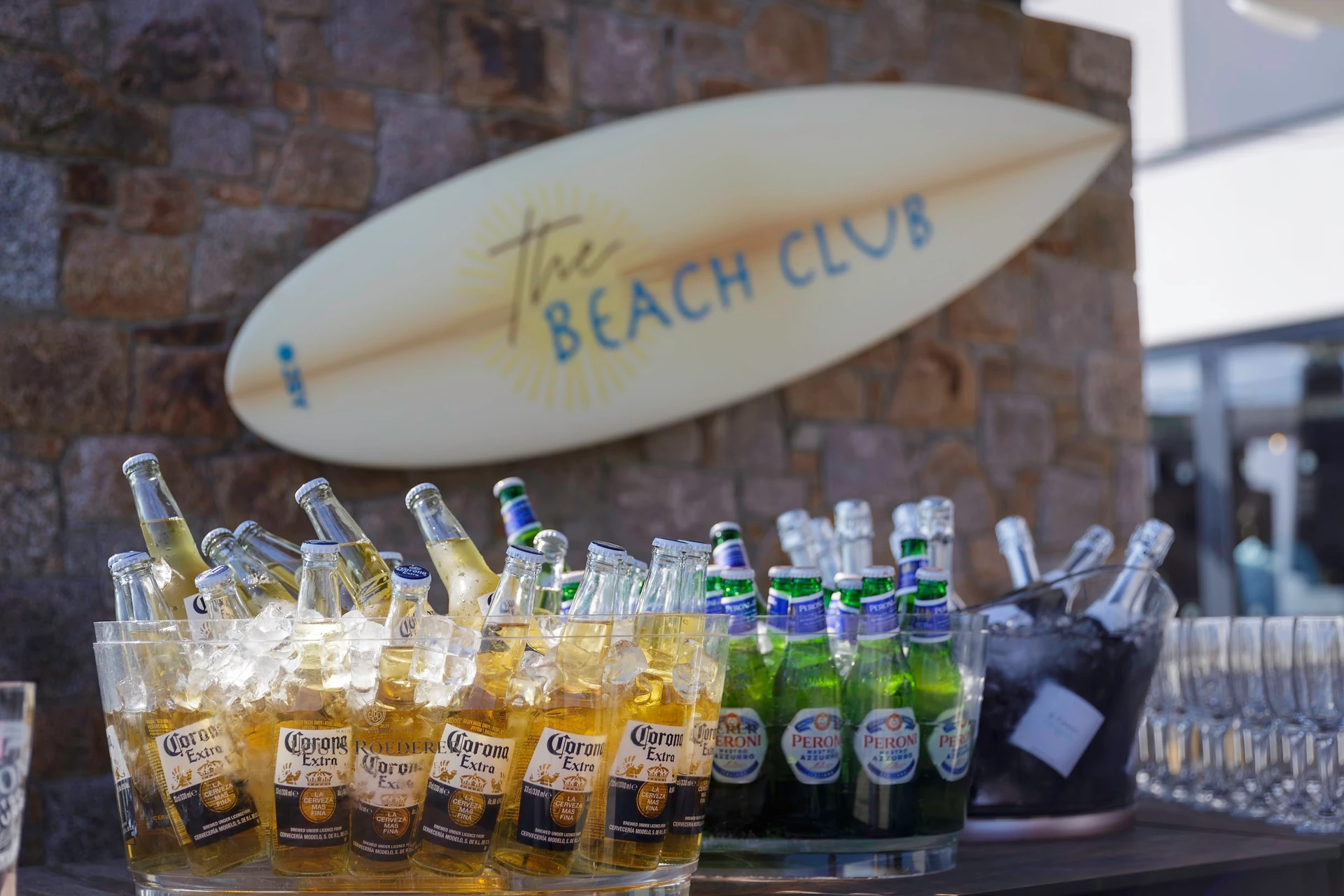 A lively beach bar scene features a stone wall with a "Sailaja" surfboard, a table with 12 clear plastic bottles of "Sailaja" soda, and a bucket of ice-cold drinks. The arrangement suggests a casual, relaxed atmosphere, possibly for a beach party or gathering.