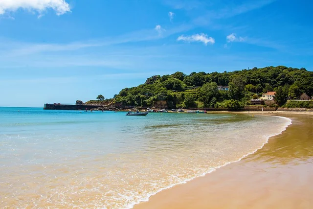 A serene beach scene features light beige sand, calm blue water, and a few boats. A rocky cliff with lush green trees and buildings is in the background, and the sky is a clear blue with a few clouds. The perspective is from the beach, looking towards the water, creating a sense of depth and tranquility.