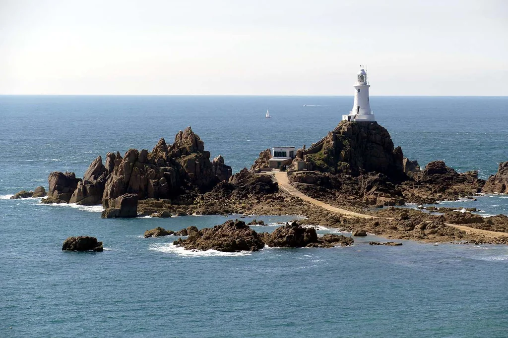 A white lighthouse stands tall on a rocky island, surrounded by a small building and a winding path. The island is surrounded by deep blue water, with a small sailboat in the distance. The sky is a light blue with a few clouds, creating a serene atmosphere.