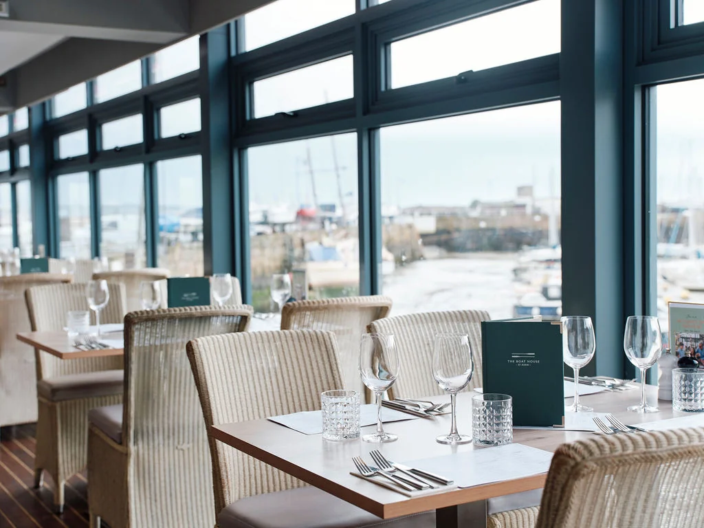 The image shows a modern restaurant with large windows offering a view of the harbor. The restaurant features white tables with black napkins and silverware, and is furnished with beige chairs. A green menu is displayed on the table, and the restaurant is well-lit by natural light. The scene is serene and inviting, with a harmonious blend of modern design and natural beauty.