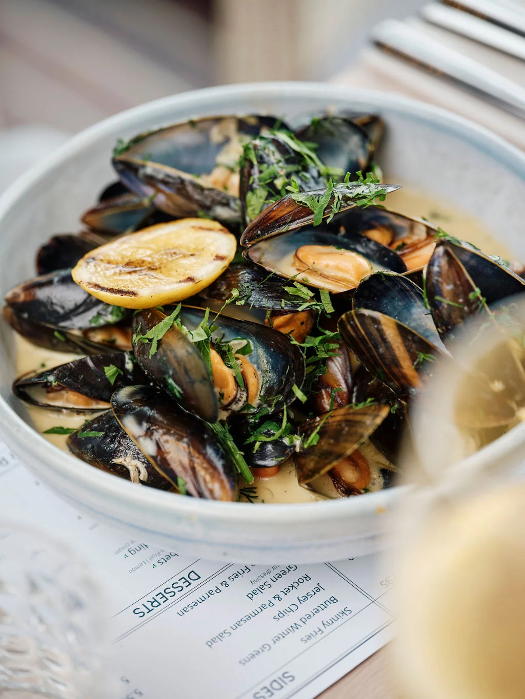 A white bowl filled with mussels, garnished with lemon and herbs, sits on a white tablecloth with a menu in the background. A glass of white wine is nearby, and a fork is ready for use. The scene suggests a dining experience at a restaurant named "Bouquet de la Cité."