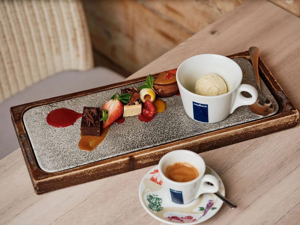 A wooden table features a rectangular tray with a chocolate cake, strawberries, ice cream, and a cup of coffee. The tray is on a wooden surface, and the coffee is on a saucer with a spoon. The scene is inviting and visually appealing.