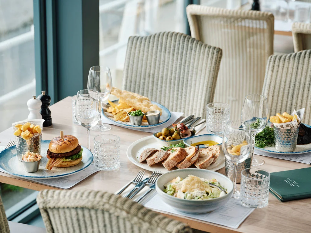 The image shows a well-arranged dining table with a blue and white tablecloth, featuring a variety of dishes and drinks. The table is set for four people, with plates of food and drinks, utensils, and condiments. The background shows a window with a view of a building, creating a serene atmosphere. The scene is visually appealing and suggests a well-prepared meal for a social gathering.
