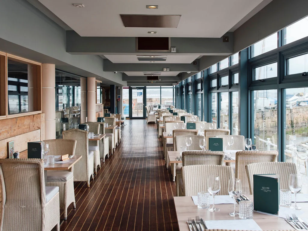 The image shows a modern, minimalist restaurant with a high ceiling, large windows, and a wooden floor. The restaurant features white chairs with green napkins and menus, a bar with a green menu, and a green bookshelf. The layout is well-organized, with tables and chairs arranged in a U-shape, and the bar and menu are centrally located. The restaurant is well-lit, creating a welcoming atmosphere.