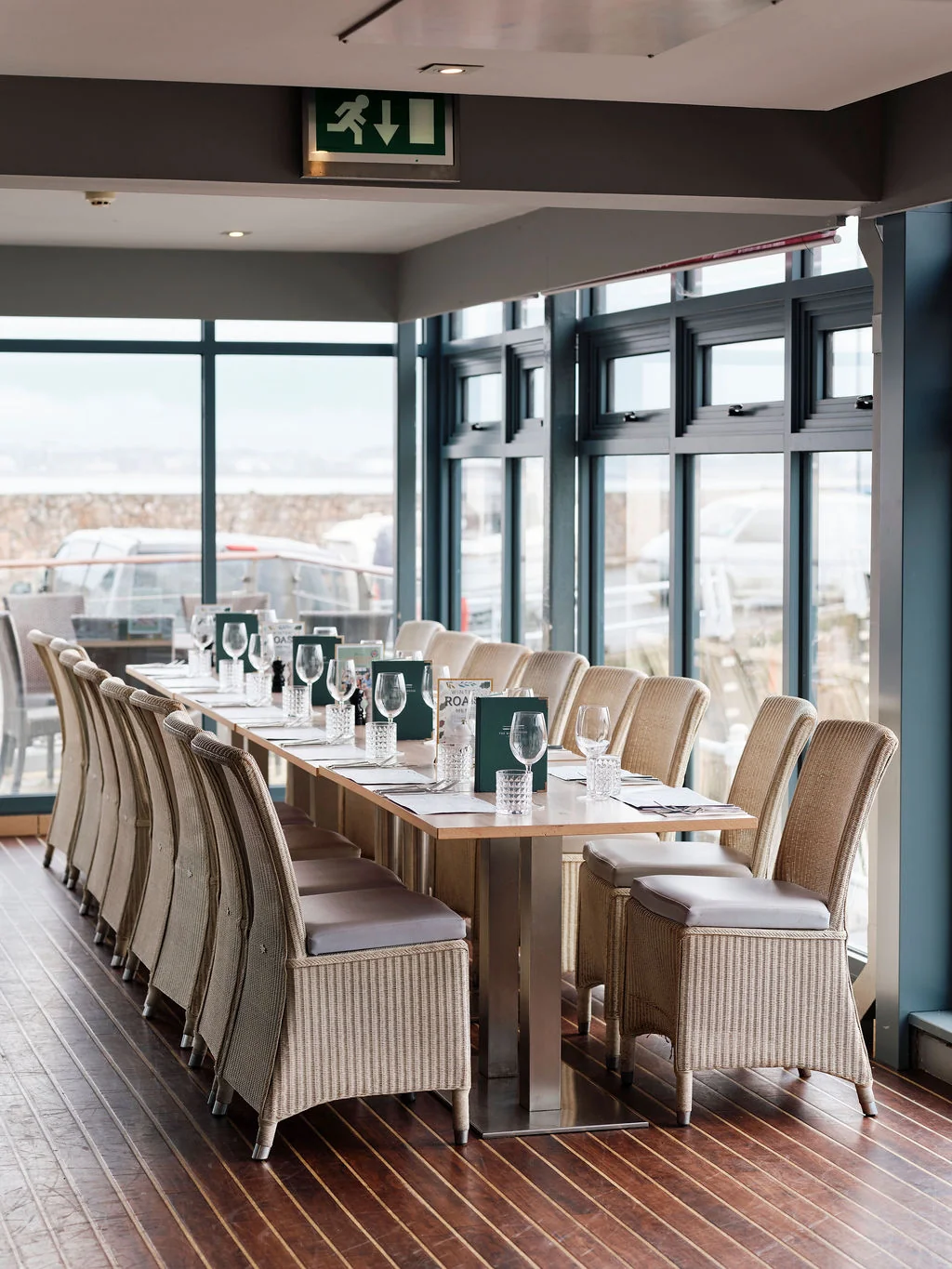 The image shows a modern restaurant with a long, white table set for eight, surrounded by beige chairs with green cushions. The table is adorned with white napkins and glasses, and the room has large windows offering a view of the cityscape. The restaurant is well-lit and has a modern aesthetic, with a green exit sign indicating the exit.