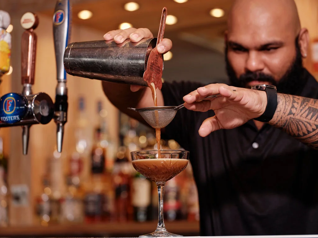 A bartender in a black shirt is pouring dark brown cocktail into a glass with a silver shaker, using a wooden bar as the backdrop. The background features various liquor bottles and shakers, creating a lively atmosphere. The bartender's focus is on the task at hand, showcasing the art of bar culture.