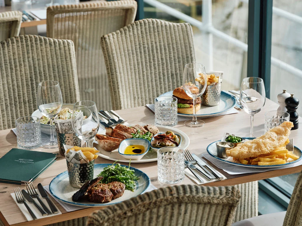 The image shows a well-arranged dining table with a variety of dishes, including a burger, fries, salad, and salad dressing. The table is set with silverware, glasses, and a black menu. The table is surrounded by wicker chairs, and the background features a window with a view of a balcony. The scene is serene and inviting, with a focus on the dining experience.