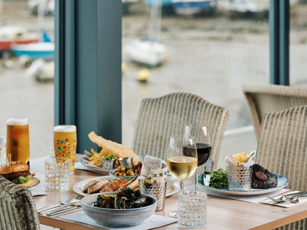 A dining table with a white tablecloth and two chairs is set for a meal overlooking a harbor with boats. The table features a plate of food, a bowl of fries, a plate of meat, a bowl of salad, and a glass of red wine. The background shows a large window with a view of the harbor. The scene is serene and inviting, with a focus on the dining experience.