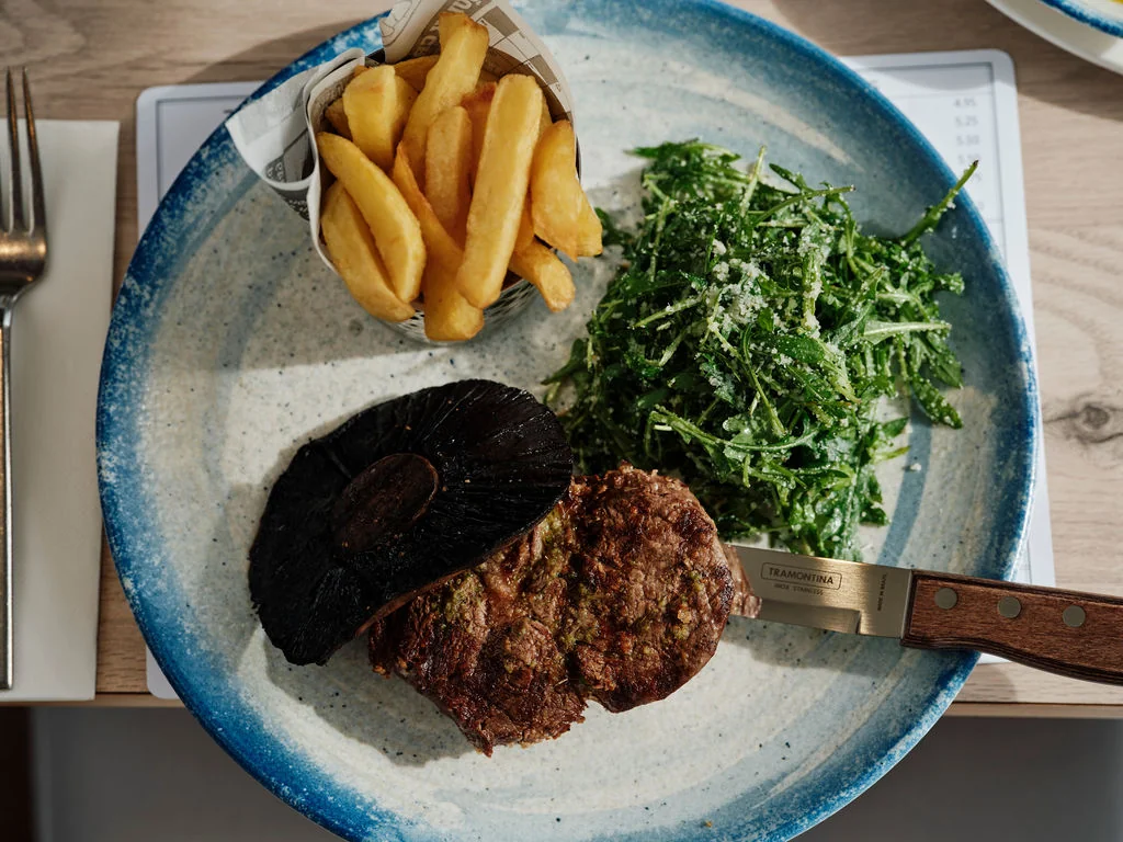 A white plate with a blue rim holds a burger, macaroni salad, and french fries, arranged on a white tablecloth. The burger has a browned meat patty, a dark brown bun, and a browned bun. The macaroni salad has green leaves, and the french fries are golden brown. A knife with a wooden handle is on the plate, ready for use. The arrangement is visually appealing and appetizing.