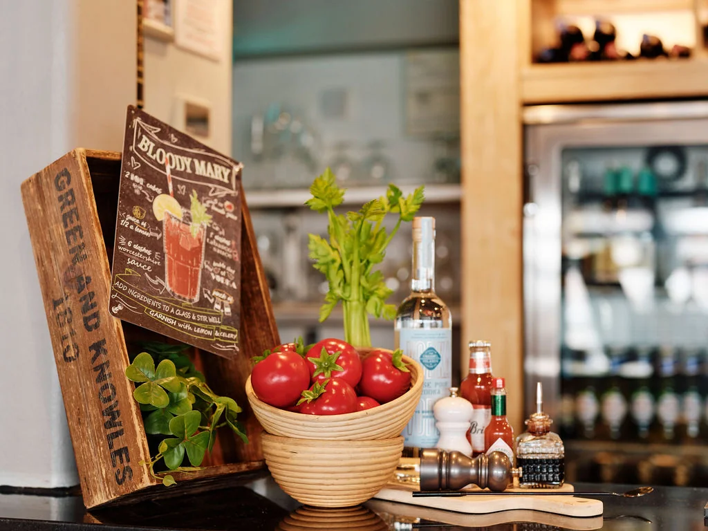 The image features a cozy bar counter with a wooden board displaying a menu and a wooden bowl filled with fresh tomatoes. A white salt shaker and pepper shaker are nearby, along with a wooden cutting board. A plant adds a touch of nature, and a wooden shelf in the background holds various bottles of alcohol. The scene is inviting and well-organized, with a focus on the bar's offerings.