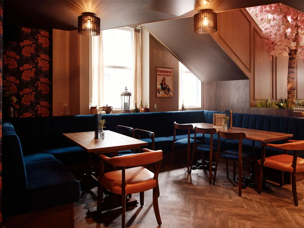 The image depicts a cosy, well-lit restaurant with a blue sofa, wooden tables, and chairs. The walls are adorned with floral wallpaper and a poster, while the ceiling features hanging lights and a black and white sign. The floor is made of light-colored wood, and the room is filled with natural light from a large window. The overall atmosphere is warm and inviting, perfect for socializing.