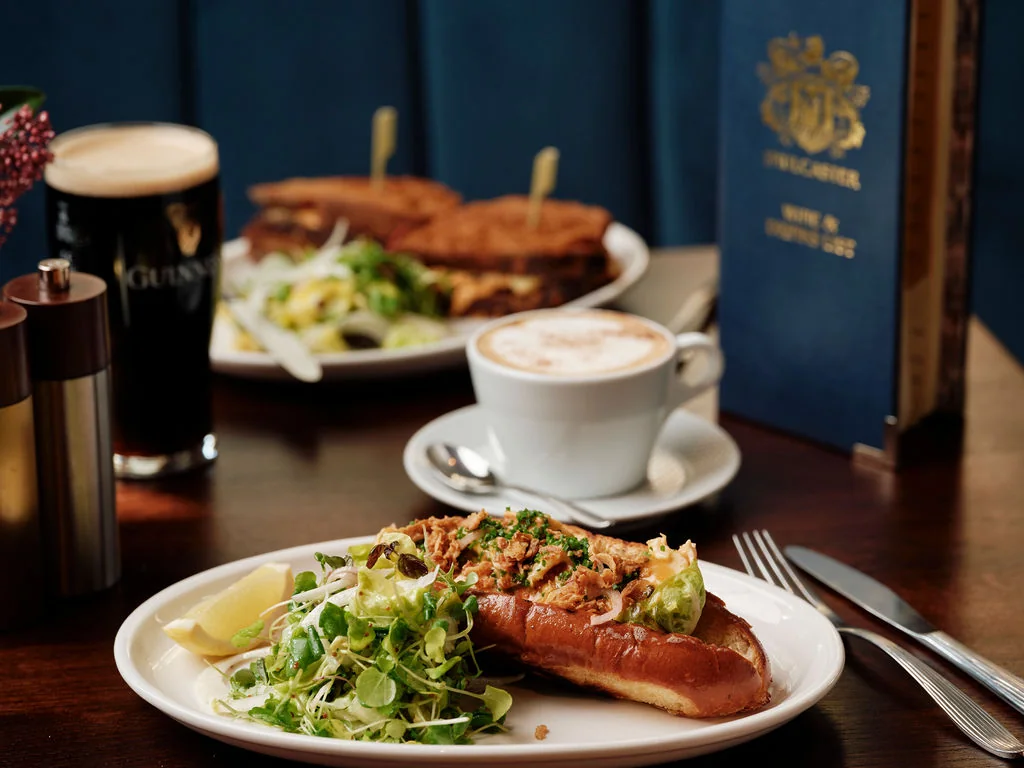 A cozy restaurant setting features a wooden table with a white plate holding a sandwich, salad, and a glass of dark beer. A silver fork and knife are nearby, and a black book with gold text is in the background. The scene is inviting and suggests a meal for two.