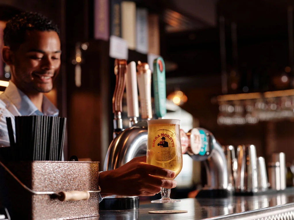 A smiling bartender in a white shirt and black hair stands behind a shiny silver bar, holding a glass of golden beer with a black logo. The bar is filled with beer taps and glasses, and a tray of black straws is nearby. The background features a well-stocked bar with various beers and glasses, creating a lively and inviting atmosphere.