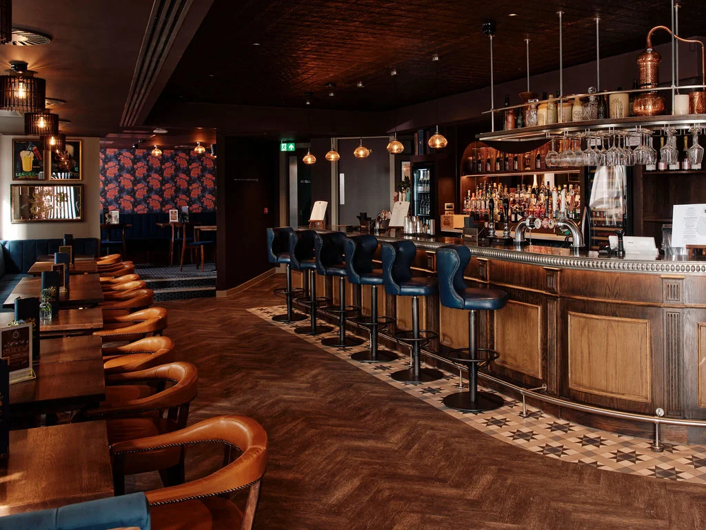 The image shows a modern, well-lit bar with a wooden bar, blue bar stools, and a red and black patterned wall. The floor is a checkered pattern, and the bar has a shelf with various bottles and glasses. The atmosphere is cosy and inviting, with hanging lights and a green exit sign.