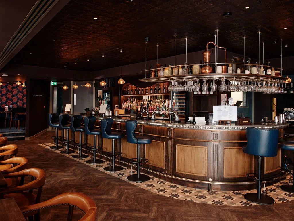The image depicts a modern, well-lit bar with a wooden bar, blue bar stools, and a variety of liquor bottles on shelves. The bar is surrounded by a black and white checkered floor, and a red and black patterned rug adds color. A man in a blue shirt and a woman in a black shirt are present, along with a bar staff member. The bar is equipped with a silver bar stool, a silver bar stool, and a silver bar stool with a glass top. The bar is illuminated by a white light fixture and a green light fixture. The overall atmosphere is inviting and sophisticated.