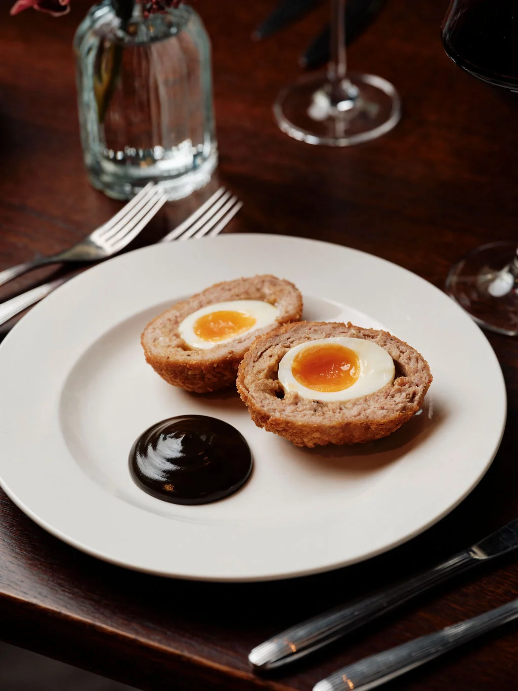 A wooden table features a white plate with two eggless hams on a bread roll, accompanied by a dark red sauce. Two silver forks and knives are on the left side, while a wine glass and vase with a flower are on the right. The scene is set for a luxurious and indulgent meal.