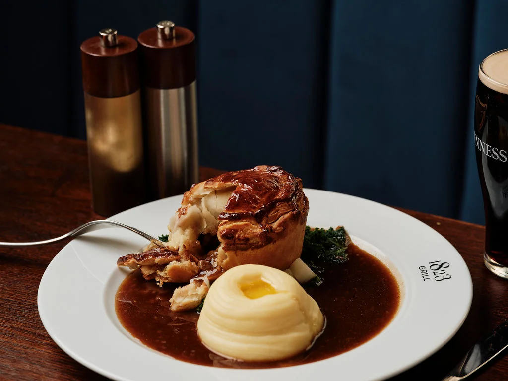 A white plate with a golden-brown roast, mashed potatoes, and a dark brown sauce is the centerpiece of a cosy dining scene. Two silver salt and pepper shakers are to the right, and a dark brown beer glass with a frothy top is on the left. The dark blue wall serves as a backdrop, creating a warm and inviting atmosphere.