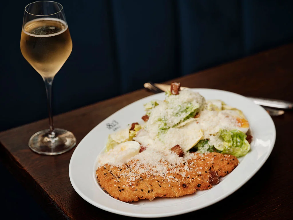 A white plate with golden-brown chicken, lettuce, and cheese sits on a dark blue table, accompanied by a glass of white wine. The chicken is garnished with black pepper and sesame seeds, while the lettuce and cheese are sprinkled with parsley. The wine glass is filled with a pale yellow liquid, creating a harmonious color palette. The scene is set against a dark blue wall, emphasizing the colors and textures of the food and drink.