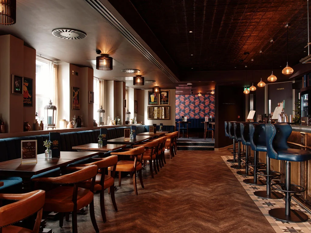 The image depicts a spacious, well-lit bar with a wooden bar, a counter, and a bar stool. The bar is adorned with a vase of flowers and a lamp, while the counter has a blue and white patterned tablecloth. The walls feature a painting, a painting with a floral design, and a large window with white curtains. The floor is covered in a patterned carpet, and the ceiling has a ceiling light fixture. The bar is equipped with a bar stool and a bar stool with a blue cushion.