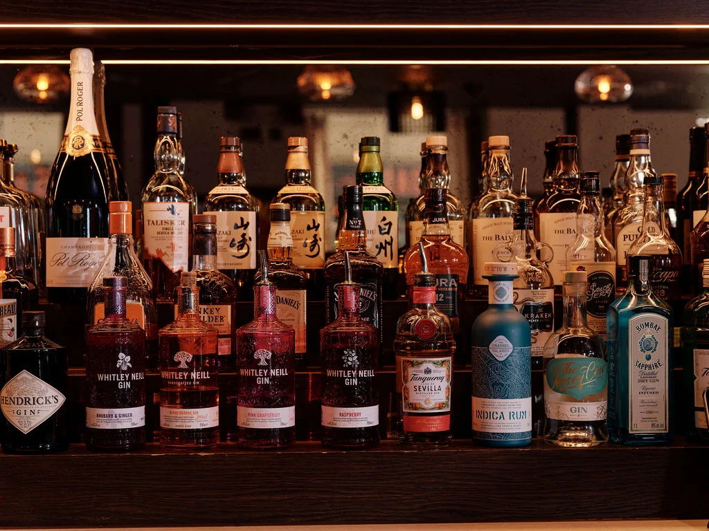 The image features a well-organized bar display with 24 distinct liquor bottles, each with unique labels and colors. The bottles are arranged in rows, with some bottles on the left and others on the right. The background is dimly lit, creating a warm atmosphere. The arrangement suggests a well-maintained bar, ready to serve guests.