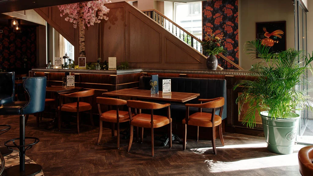 The image depicts a cozy, well-lit restaurant with a warm, inviting atmosphere. The wooden floor and walls are adorned with floral wallpaper, and a wooden bar with a white sign is present. Tables with orange leather seats and wooden chairs are scattered throughout the space, with a blue trash can nearby. A potted plant and a pink flower add color to the scene. The restaurant is well-organized and welcoming, with a focus on comfort and style.