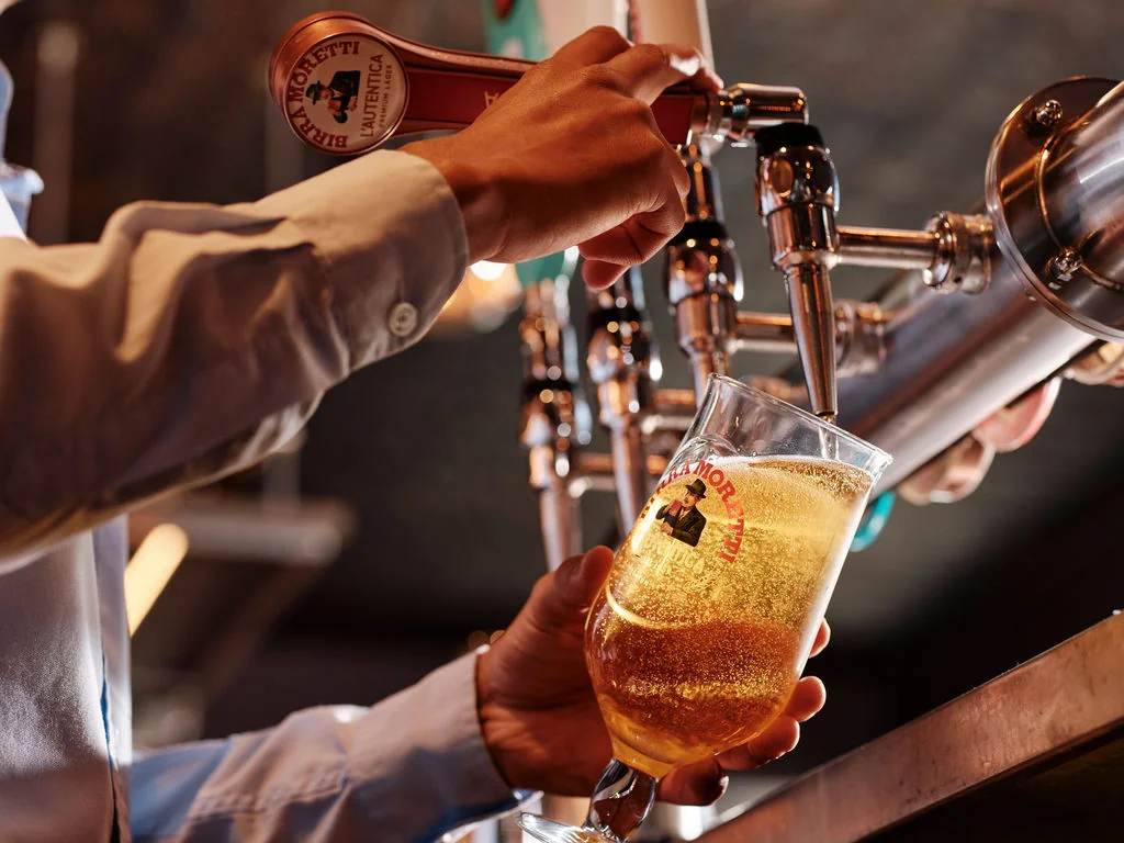 A person in a blue shirt is pouring beer into a glass at a bar, using a silver tap with a red label. The glass is filled with a golden liquid, and the background is blurred, emphasizing the action. The image captures a moment of enjoyment and relaxation in a social setting.