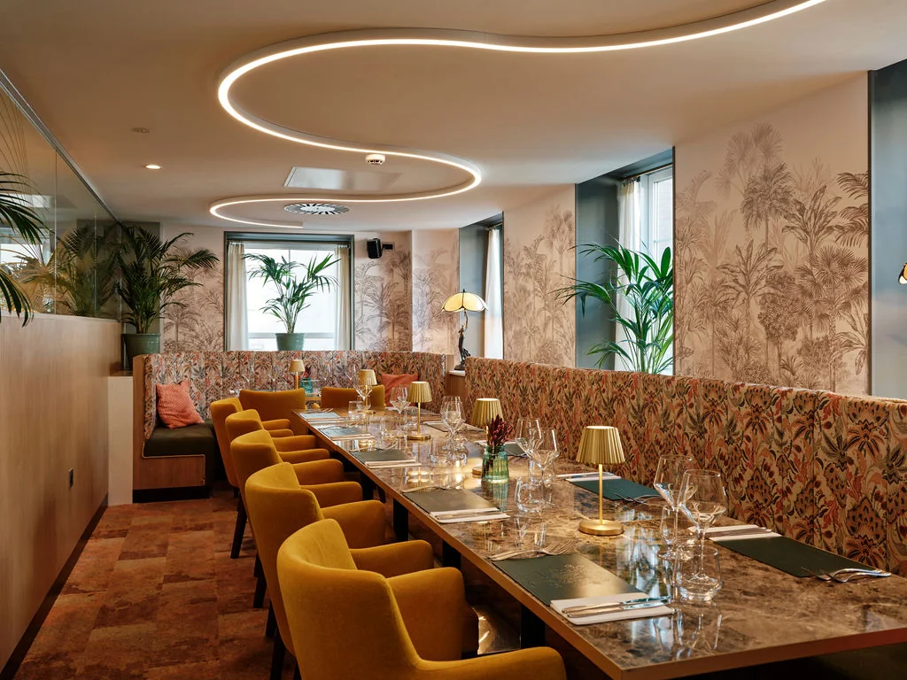The image shows a modern, elegant restaurant with a neutral color palette, featuring a long table with a white marble top, surrounded by yellow chairs and a floral-patterned booth. The tables are set with silverware, wine glasses, and a vase of flowers. The walls are adorned with floral wallpaper, and the ceiling has circular lights. The floor is made of wood, and the restaurant is well-lit, creating a warm and inviting atmosphere.