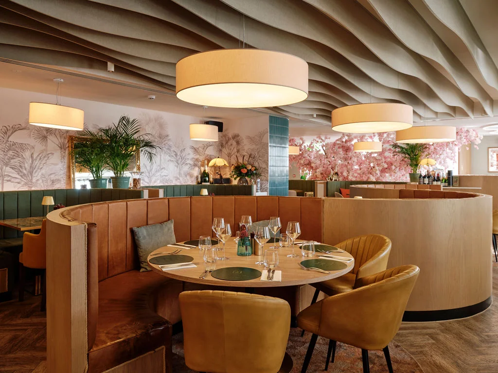 The image depicts a modern, elegant restaurant with a curved wooden ceiling, white walls, and a wooden floor. The walls feature a floral pattern, and the ceiling has hanging lights. The restaurant has a wooden bar with a green bench, and the tables are set with green napkins, white plates, and green glasses. The seating arrangement includes yellow chairs with green cushions, and the tables are surrounded by green and yellow plants. The restaurant is well-lit and has a cozy atmosphere.