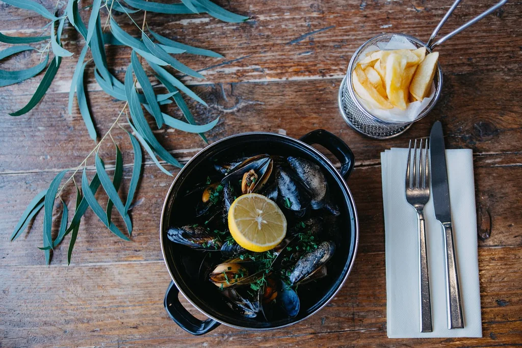 A wooden table features a black cast iron skillet with mussels and a lemon slice, a white napkin, a fork, knife, and a glass of fries with a straw. A green leaf adds a touch of nature. The scene is inviting and visually appealing.