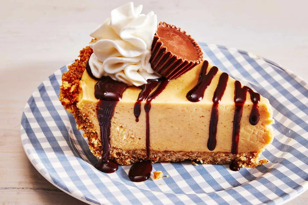 A slice of cheesecake with a crumbly crust, a creamy filling, and a drizzle of dark chocolate sauce is served on a blue and white checkered plate atop a wooden table. The scene is visually appealing and inviting, with the cheesecake as the star.