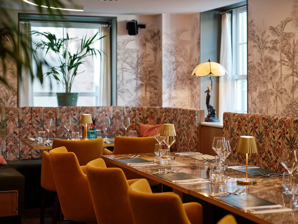 The image features a modern, elegant restaurant with a marble table, six orange chairs, and a white tablecloth. The walls are adorned with floral wallpaper and a black and white vase with a bird statue. A large window with white curtains allows natural light to illuminate the space. A lamp with a yellow shade and a plant add a touch of nature. The arrangement of objects creates a harmonious and inviting atmosphere.