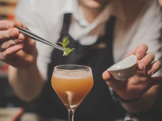 A person in a white shirt and black apron is preparing a cocktail at a table, using chopsticks to stir a garnish. The cocktail is in a tall glass with a stem, and a small spoon is nearby. The background is blurred, emphasizing the focus on the person and their cocktail-making process.