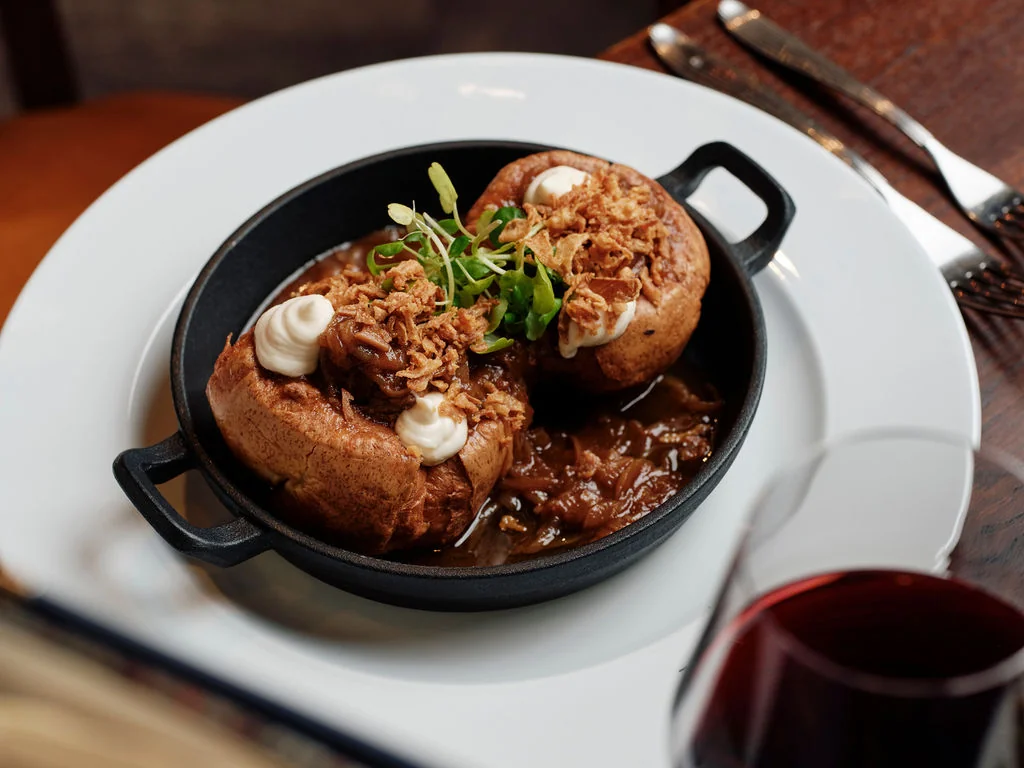 A black cast-iron skillet holds a hearty dish with brown meat, white sauce, and green garnish, accompanied by a glass of red wine on a white plate atop a wooden table. The scene is visually appealing and suggests a meal enjoyed in a cozy setting.
