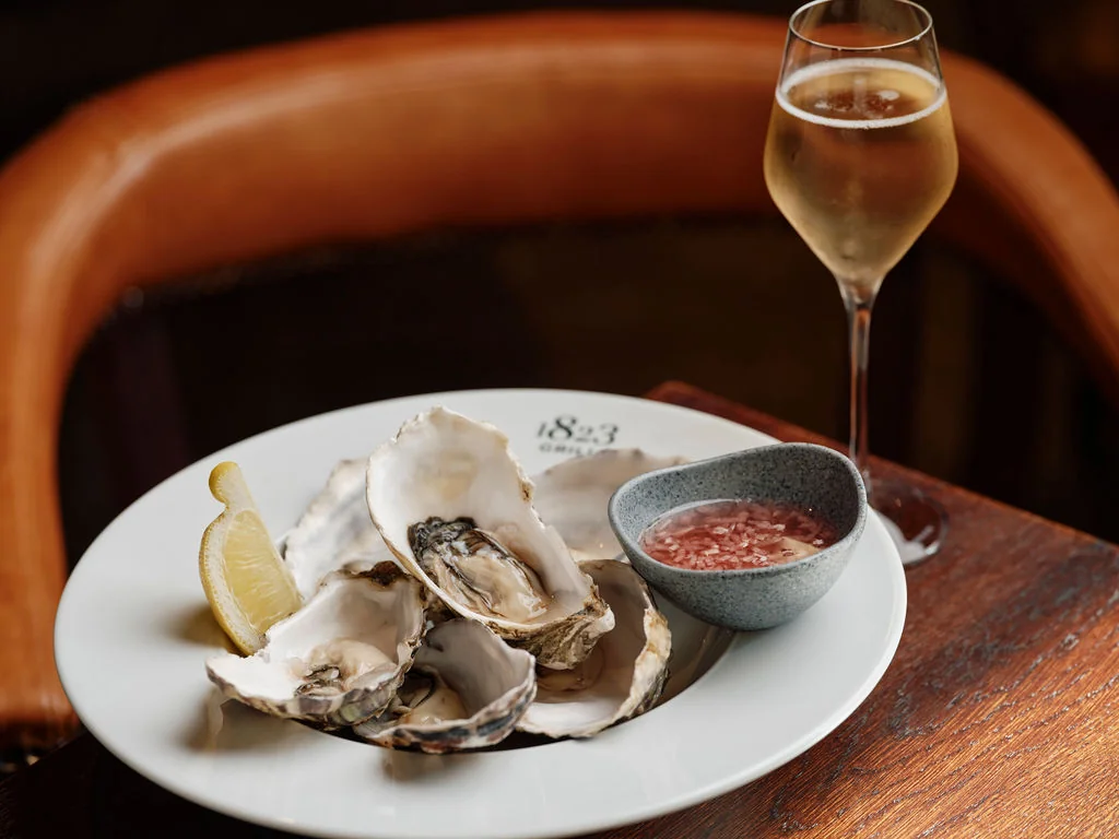 A wooden table features a white plate with oysters, a lemon wedge, and a small bowl of red sauce. A tall glass of champagne is to the right, creating a sophisticated and indulgent atmosphere.