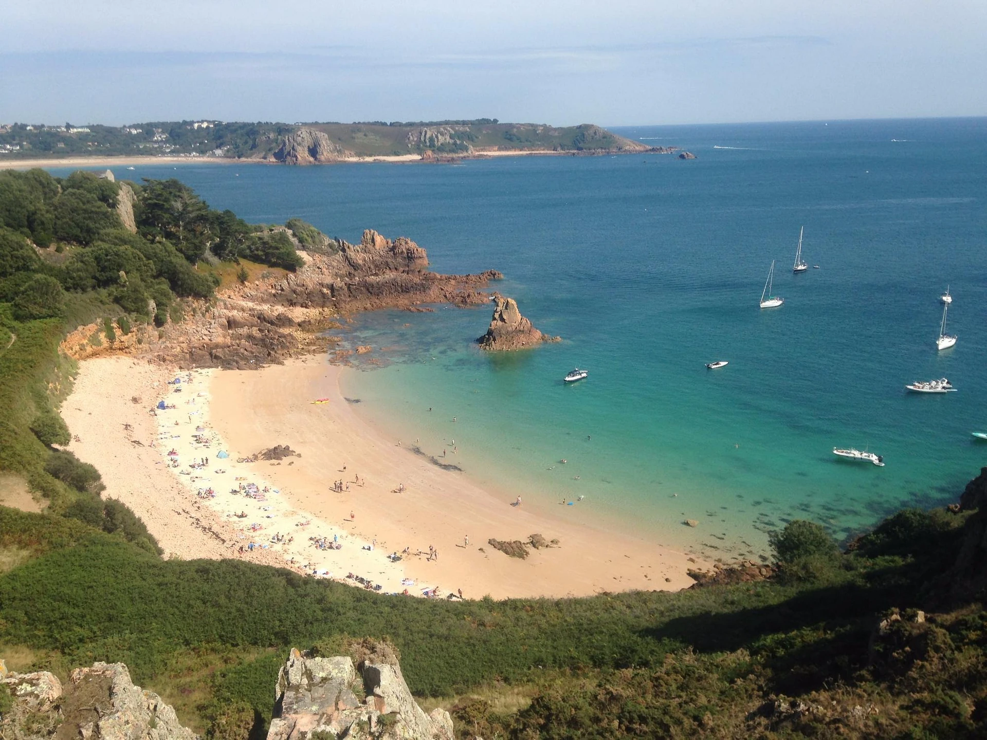 A serene beach scene features a sandy shore, clear blue water, and a few boats. The beach is surrounded by rocky cliffs and greenery, with a clear blue sky above. The image is taken from a high vantage point, offering a panoramic view of the beach, water, and cliffs. The scene is harmonious and tranquil, with no discernible text or actions.