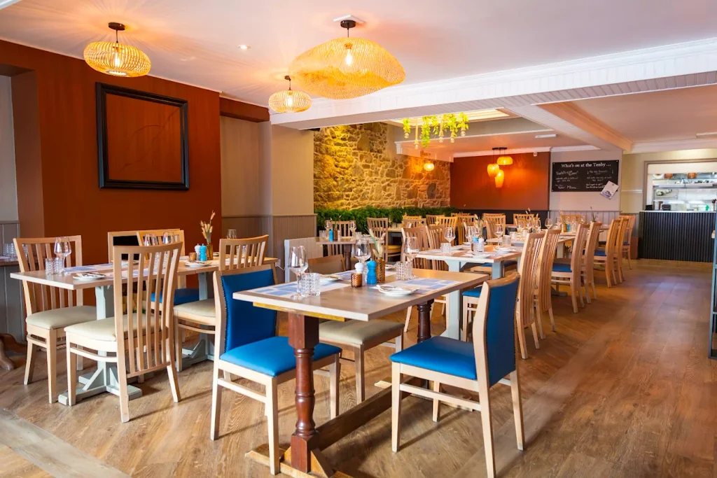The image shows a modern, well-lit restaurant with wooden floors and walls, featuring wooden chairs and tables with blue and white accents. The tables are set with white plates, silverware, and condiments. The walls are adorned with a stone wall, a blackboard menu, and hanging lights. The restaurant is empty, creating a serene atmosphere.