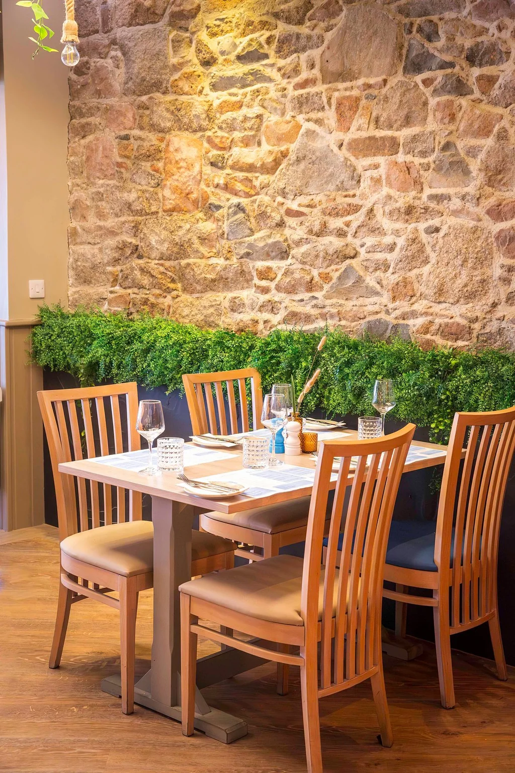 In a cozy restaurant, a wooden table with four chairs is set for four, each adorned with a white tablecloth and wine glasses. A blue vase with a flower adds color. The stone wall behind the table is adorned with green plants, creating a rustic atmosphere. The wooden floor and chandelier contribute to the warm and inviting ambiance.