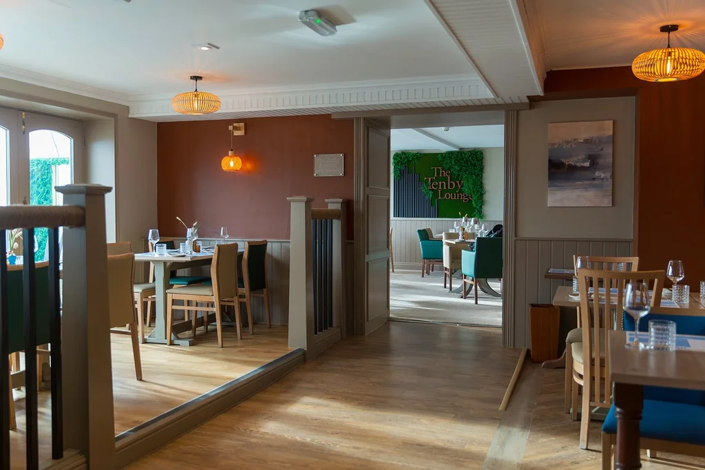 The image shows a serene, well-lit restaurant with warm wooden floors and a warm red wall. The walls are adorned with paintings and a green sign with the words "The Seaside". The restaurant has six tables with white tablecloths and silverware, and is equipped with hanging lights. The entrance is through a doorway with a wooden door and a window with a view of the sea. The restaurant is ready to welcome guests and offers a tranquil atmosphere.